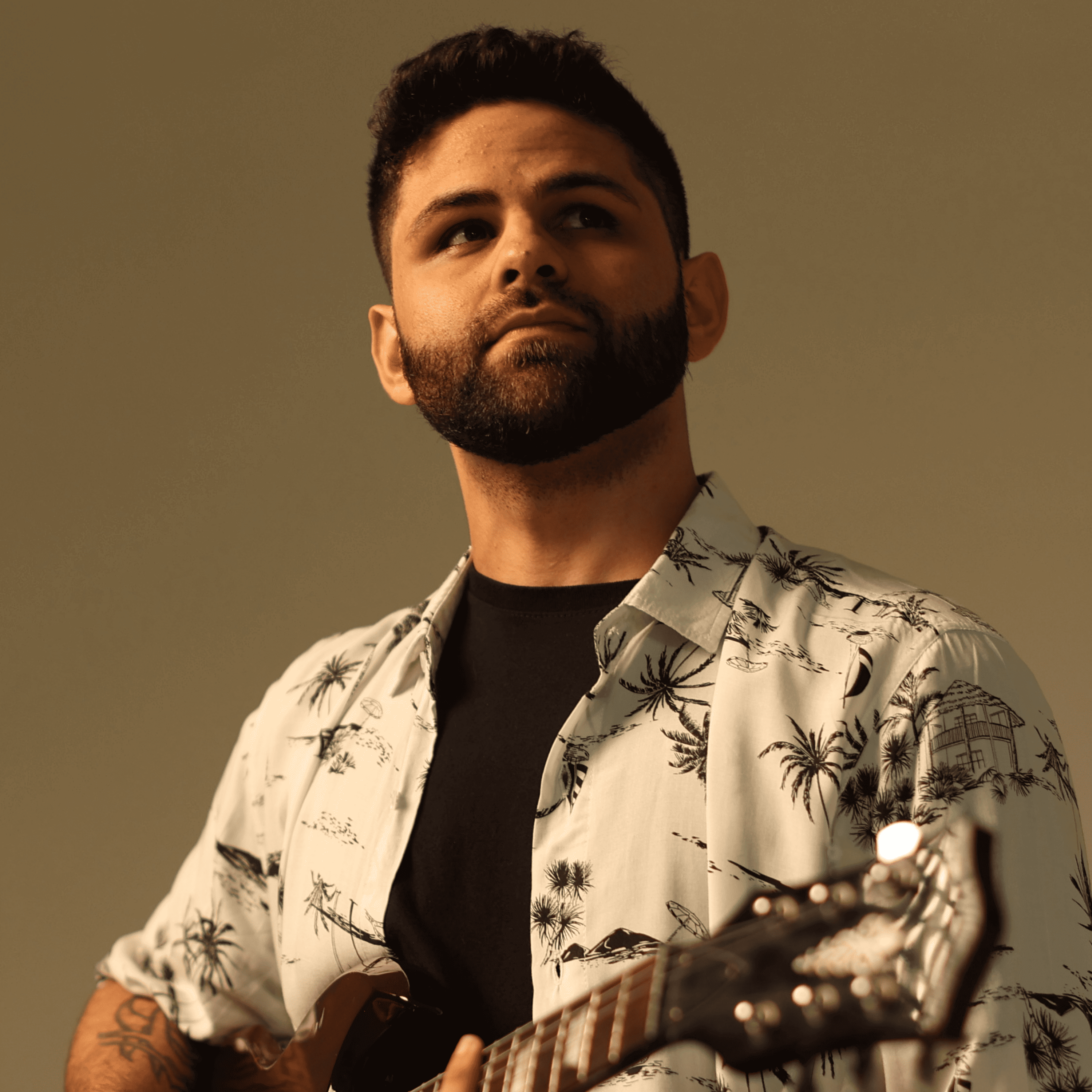 Músico Aquino, homem de barba, em perfil, sorrindo, usando camiseta estampada branca, com folhagem ao fundo.